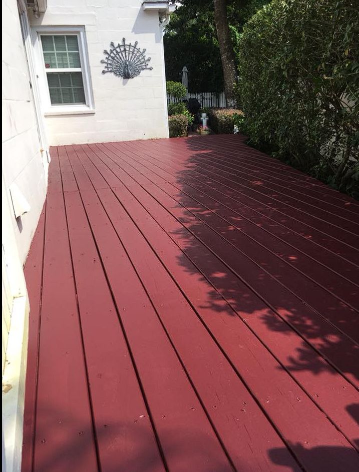 Freshly painted red wooden deck adjacent to a white house with a decorative wall ornament, surrounded by green hedges